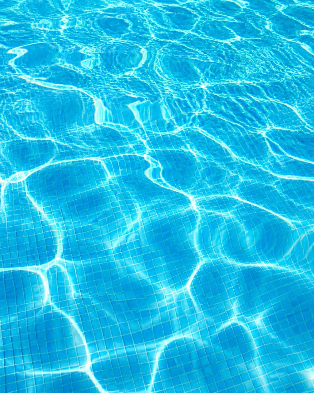 Crystal-clear turquoise pool water rippling over bright blue mosaic tiles in full sunlight, showing perfectly balanced debris-free water